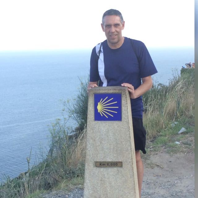 don Juan, con camisaeta azul y pantalones cortos negros, y una sonrisa de orgullo, tocando la señalización territorial del kilómetro cero coma cero cero cero del Camino de SAntiago.